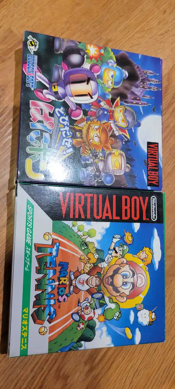 Two Virtual Boy video game boxes are stacked on a wooden surface. The top box features Hudson Soft's 'Bomberman' game with…