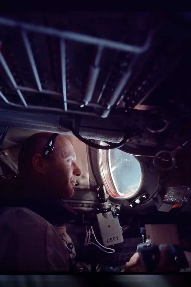 A man in a cockpit, likely an astronaut, looks out a circular window at a bright sky. The interior is dimly lit with visible…