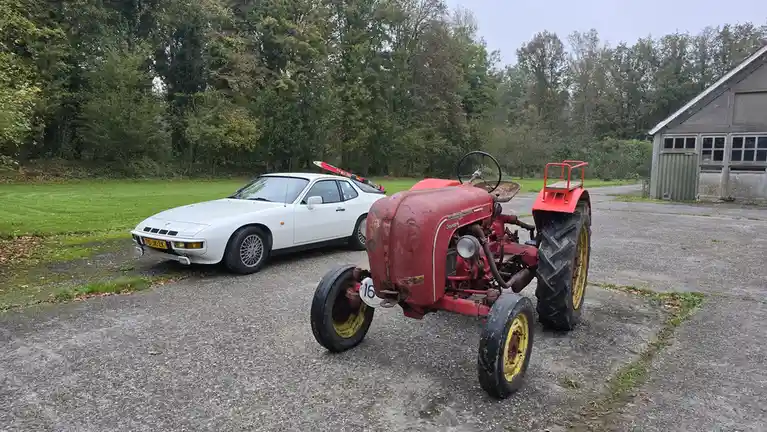 A white Porsche 924 sports car with skis mounted on its roof is parked next to a red, rusty, vintage Porsche Diesel tractor…