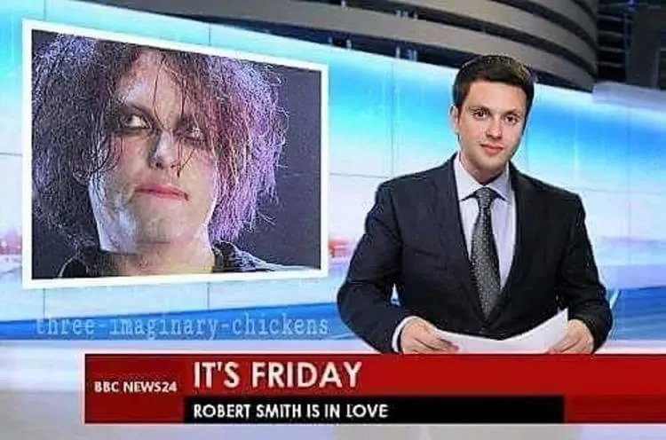 A news anchor in a suit sits at a desk with a large screen behind him showing a close-up of Robert Smith with dark makeup…