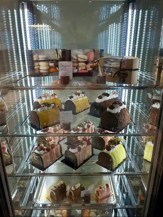 A glass display case filled with multiple tiers of assorted pastries and cakes, including macarons and rolled cakes with…