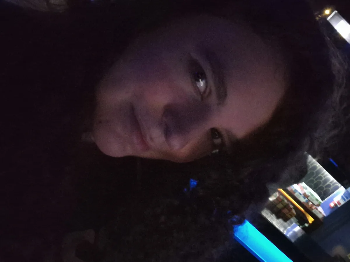A close-up, low-angle selfie of a person with dark curly hair, looking directly at the camera with a tired but slightly…