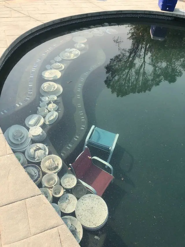 An overhead view of a swimming pool filled with numerous plates, bowls, and a chair, arranged in a curved line along the…