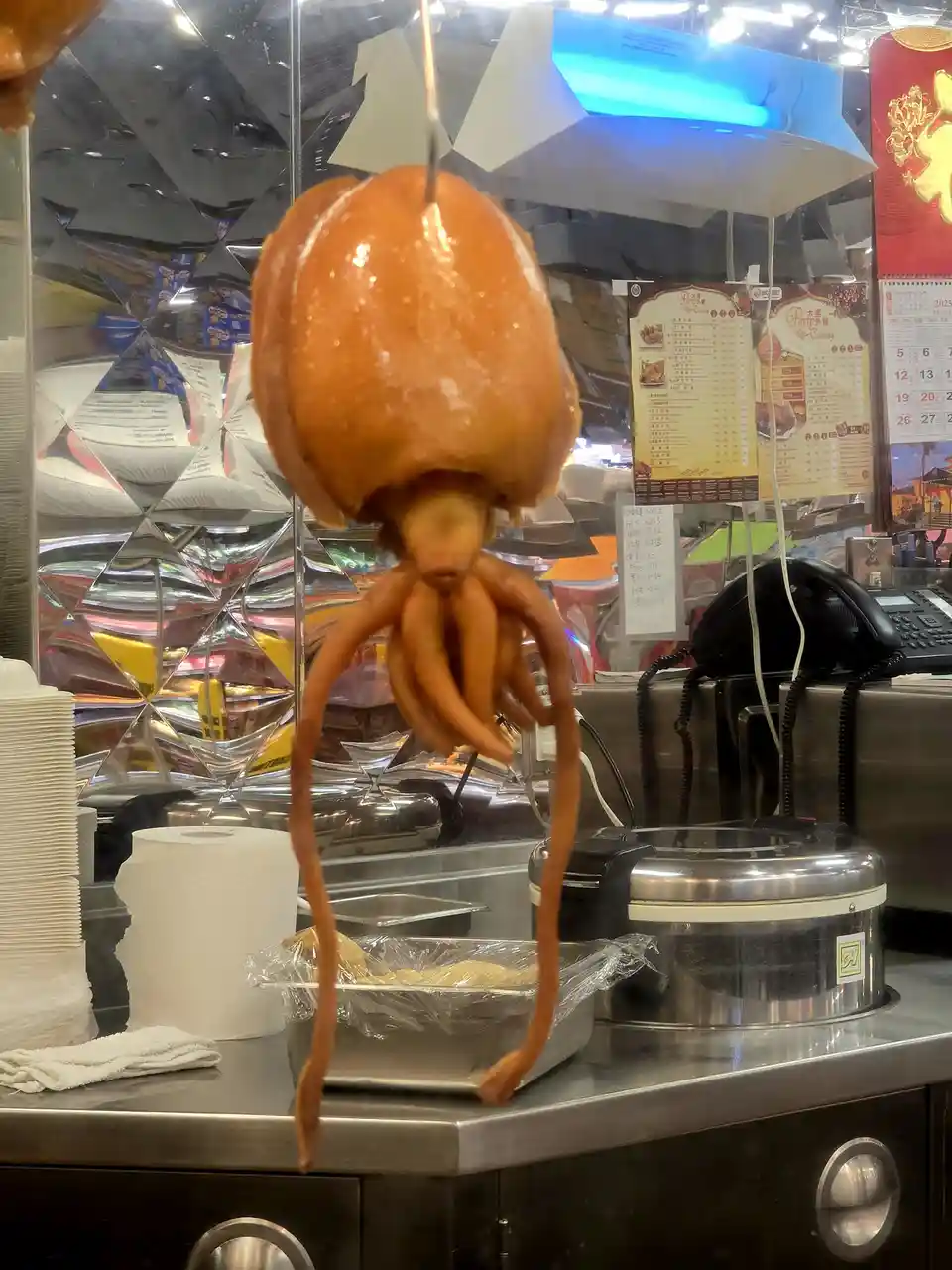 a roasted duck is hanging from a hook in a food stall, but its body has been digitally altered to have squid tentacles…