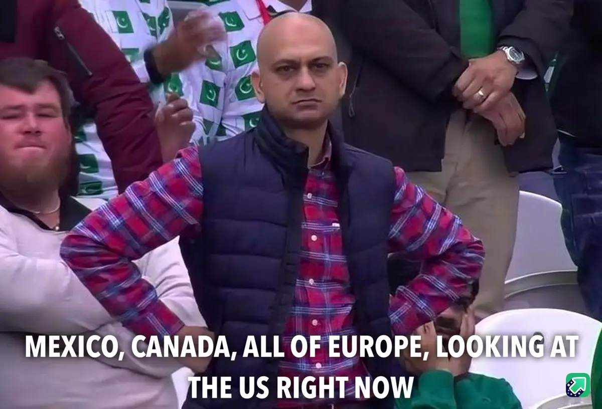 A bald man in a red plaid shirt and black vest stands with hands on hips in a stadium crowd, looking intensely forward…
