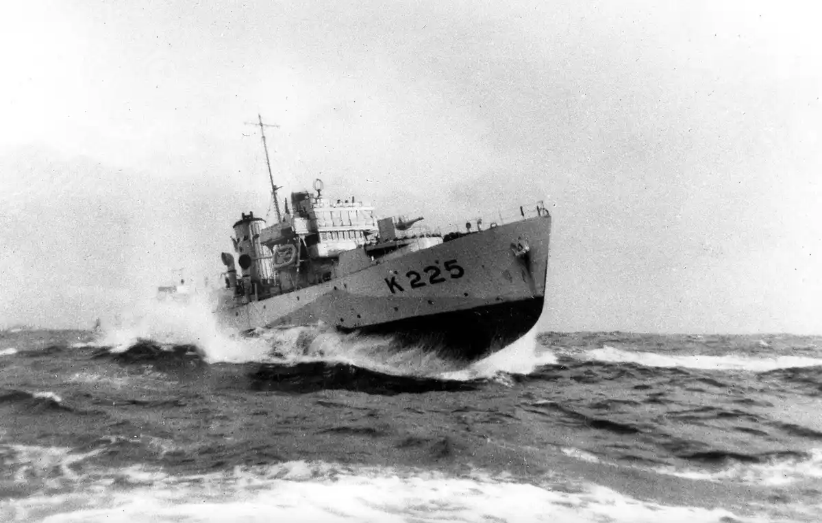 A black and white historical photograph shows a naval vessel, marked with the hull number K 225, cutting through rough ocean…