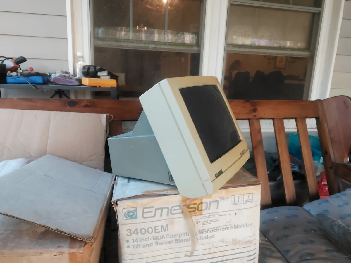 An old beige Emerson 3400EM 14-inch monochrome computer monitor sits tilted on top of its original cardboard box, which is…