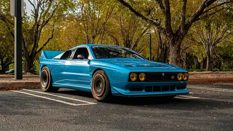 A bright blue Alfa Romeo Alfetta GTV6 is parked in a tree-lined parking lot, its distinctive quad headlights and gold-rimmed…