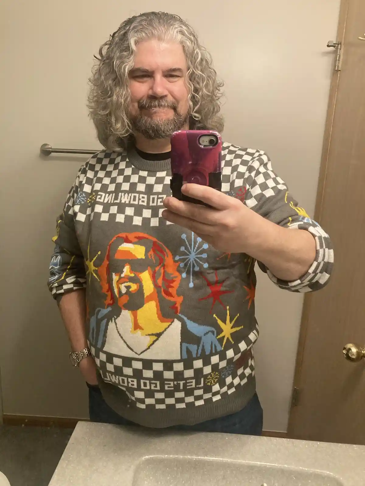 A man with long gray curly hair and a beard takes a mirror selfie in a bathroom while wearing a very colorful, patterned…