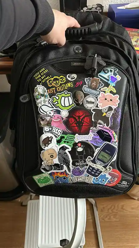 A black backpack is covered in dozens of colorful, overlapping stickers featuring anime, gaming, internet culture, and tech…