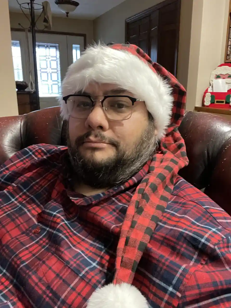 a man with a beard and glasses sits in a brown leather armchair wearing a red and black plaid shirt and a santa hat with…