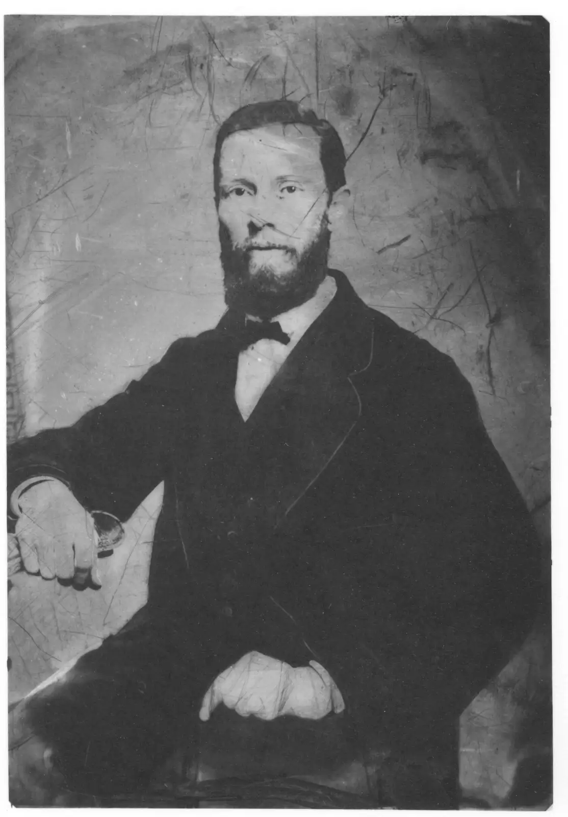A vintage black-and-white portrait of a bearded man in a formal suit and bow tie, seated and looking directly at the camera…