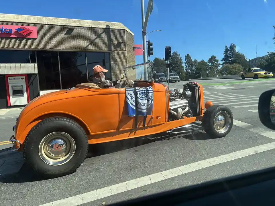 An orange hot rod with an exposed engine and two dogs in the backseat is stopped at a traffic light near a Bank of America…
