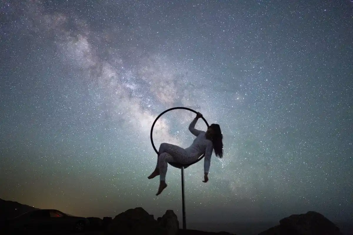 A person in pajamas is silhouetted against a starry night sky with the Milky Way visible, hanging from a hoop on a pole. The…