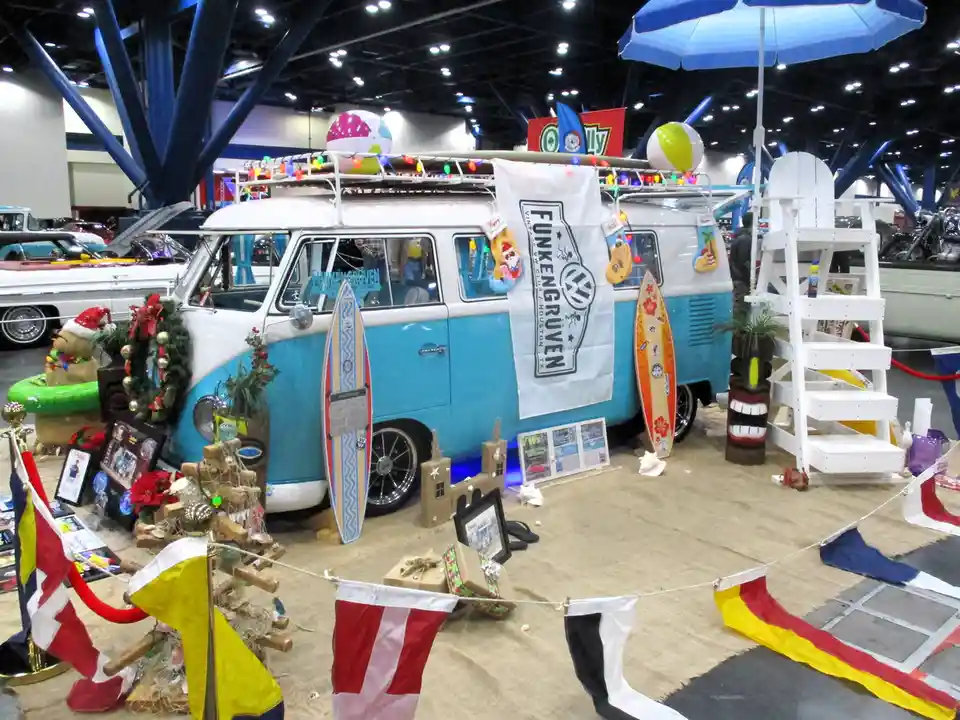 A blue and white vintage Volkswagen van is displayed at an indoor car show, decorated with surfboards, beach balls, tiki…