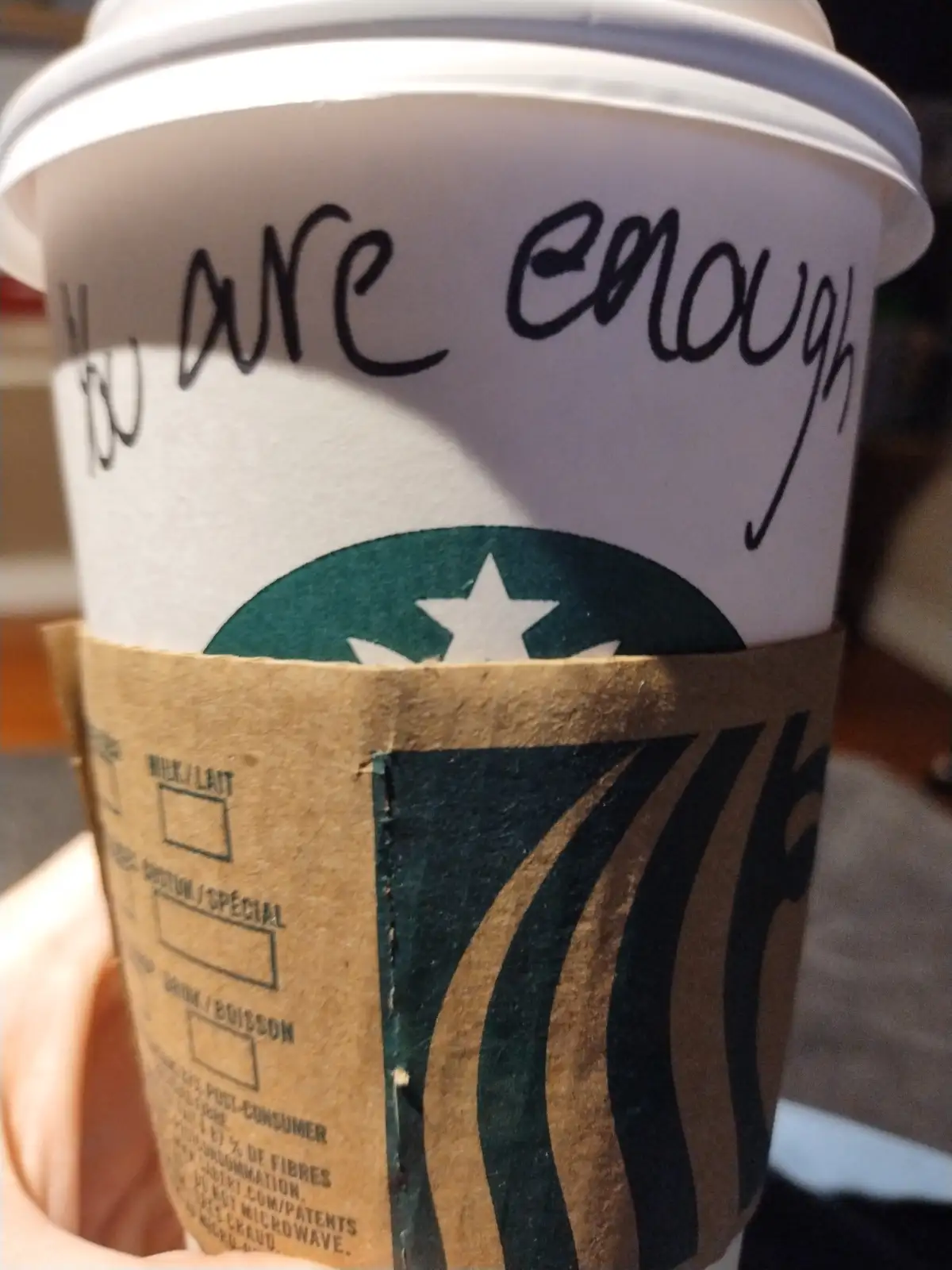A close-up photo shows a Starbucks coffee cup with a handwritten message in black marker that reads 'you are enough' above…