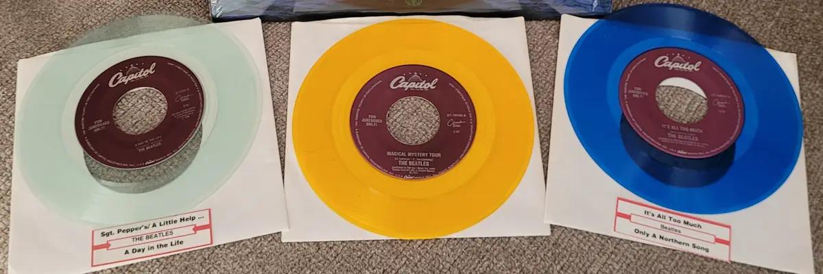 Three colorful vinyl records by The Beatles are displayed on white paper, each with a Capitol label and a red sticker below…