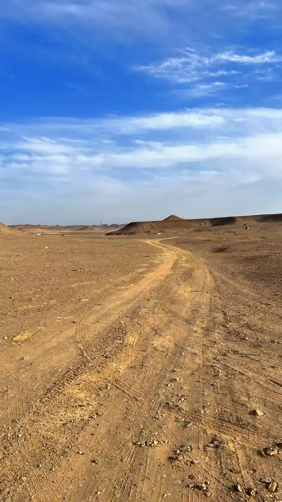 A wide-angle photo shows a long, winding dirt road stretching through a barren, arid landscape under a vast blue sky with…