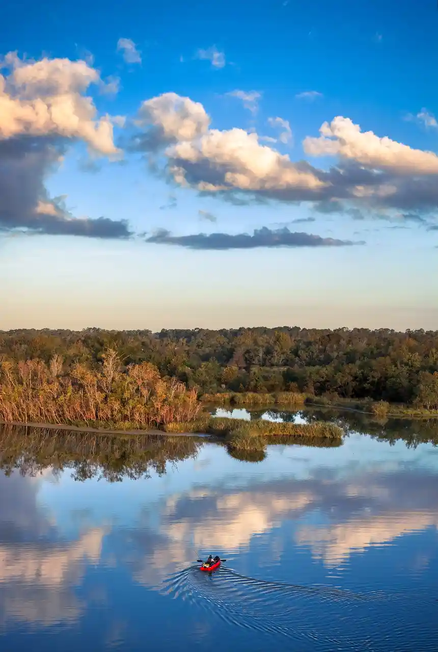 A serene landscape photo shows a small red kayak with one person paddling across a calm, reflective body of water. The sky…