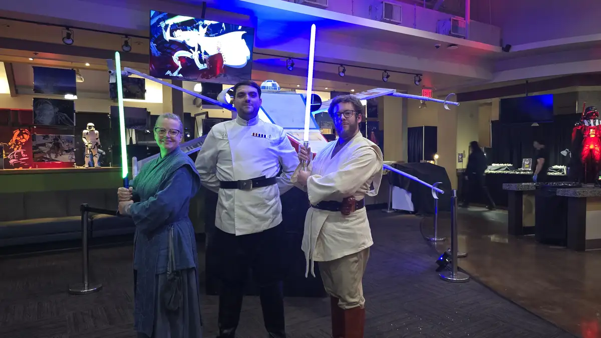 Three people dressed as Star Wars characters pose indoors holding glowing lightsabers; one wears a green robe, another a…