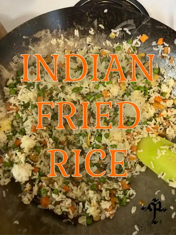 A close-up photo of a wok containing rice mixed with peas, diced carrots, and other ingredients, with a green spatula…