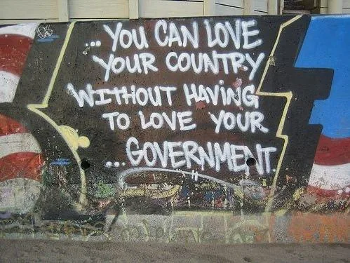 A graffiti-covered wall features bold white text stating, '... YOU CAN LOVE YOUR COUNTRY WITHOUT HAVING TO LOVE YOUR…