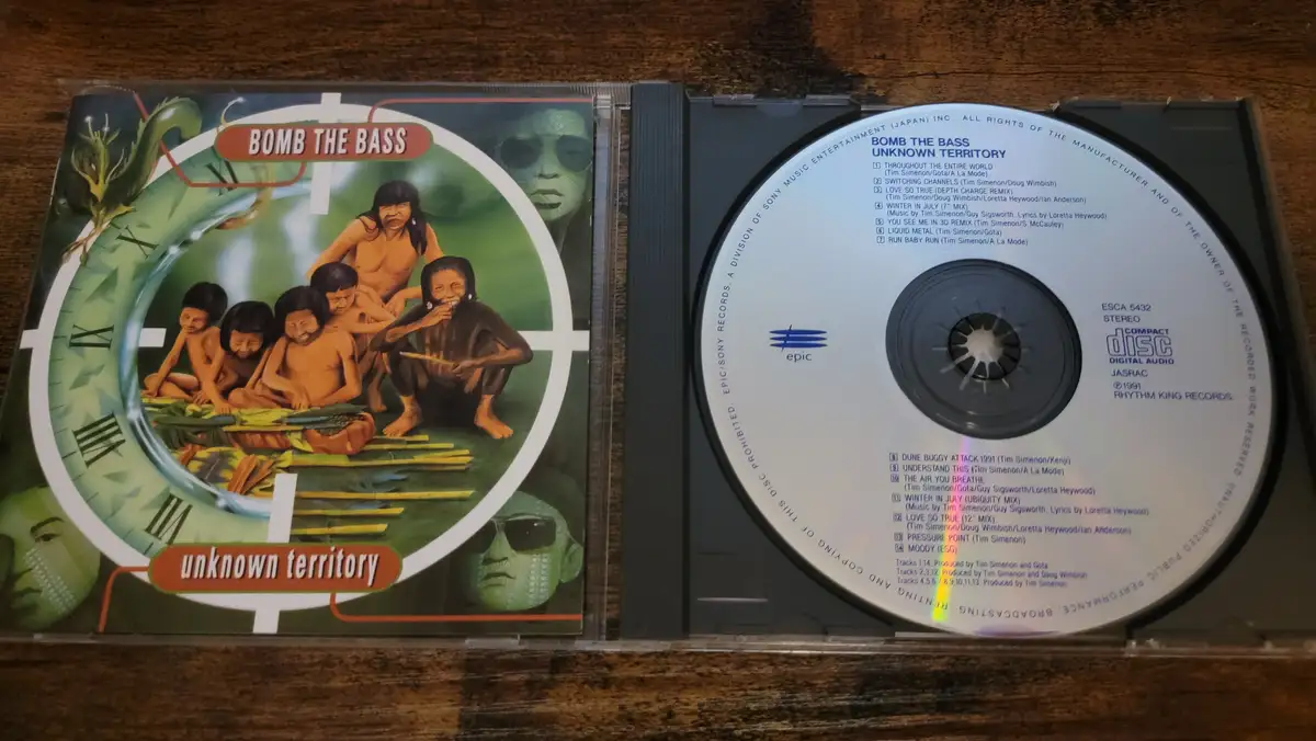 An open CD case displays the album 'Bomb The Bass - Unknown Territory' with its cover art featuring a group of people inside…
