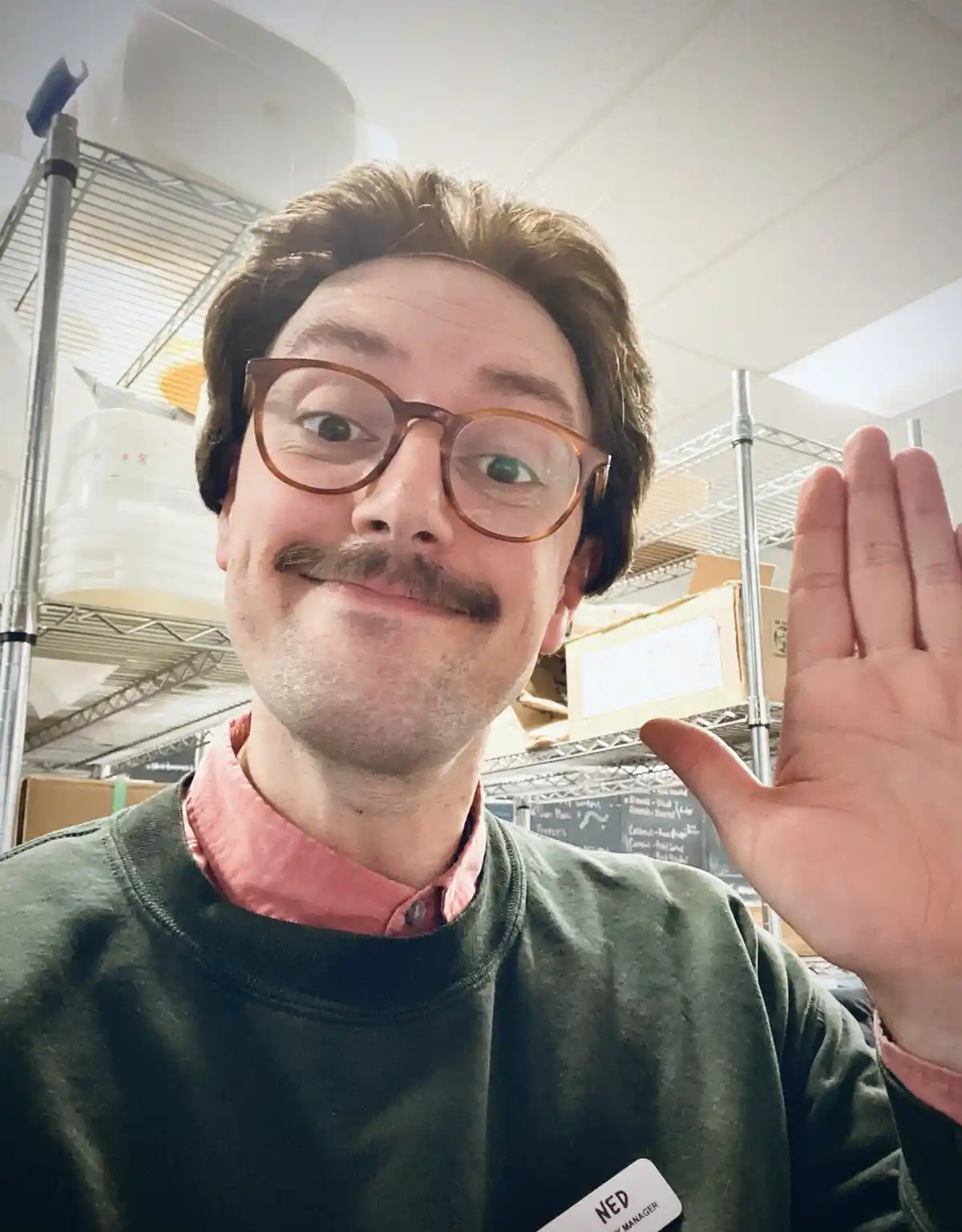 A man with glasses and a mustache, wearing a green sweatshirt over a pink collared shirt and a name tag that reads 'NED…