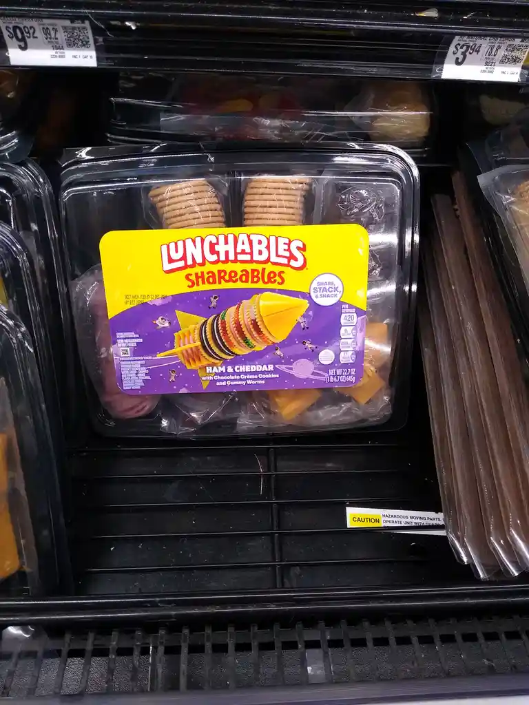 A supermarket shelf displays a Lunchables Shareables package labeled 'Ham & Cheddar with Chocolate Crème Cookies and Gummy…