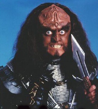 A close-up of a menacing, alien-like creature with dark skin, long black hair, and a scowling expression, holding a sword…