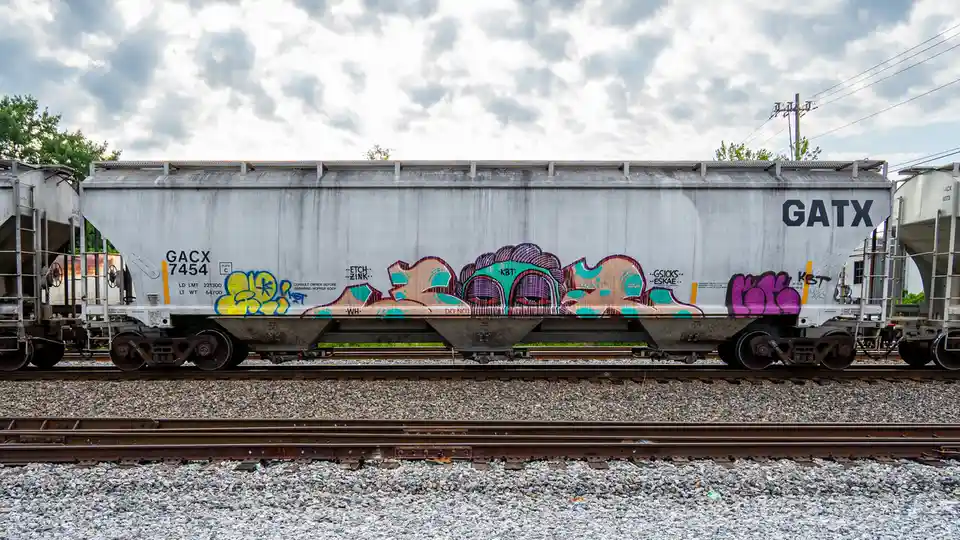 A side view of a silver GATX hopper railcar, number 7454, covered in colorful graffiti art, sits on railway tracks under a…