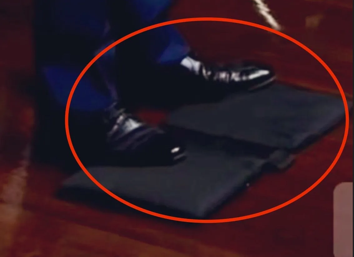 A close-up, blurry image shows a person's feet in shiny black dress shoes standing on two dark gray, folded rectangular pads…