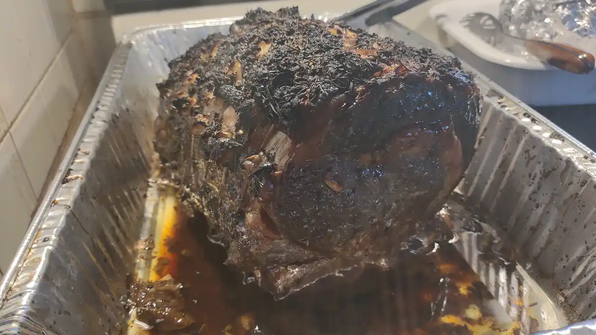 A large, heavily charred roast sits in a foil pan, its top blackened and crispy, with juices pooling below. The overlaid…