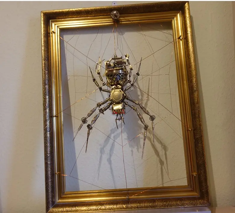 A steampunk-style spider sculpture made of brass, gears, and wires is suspended in the center of a large ornate gold picture…