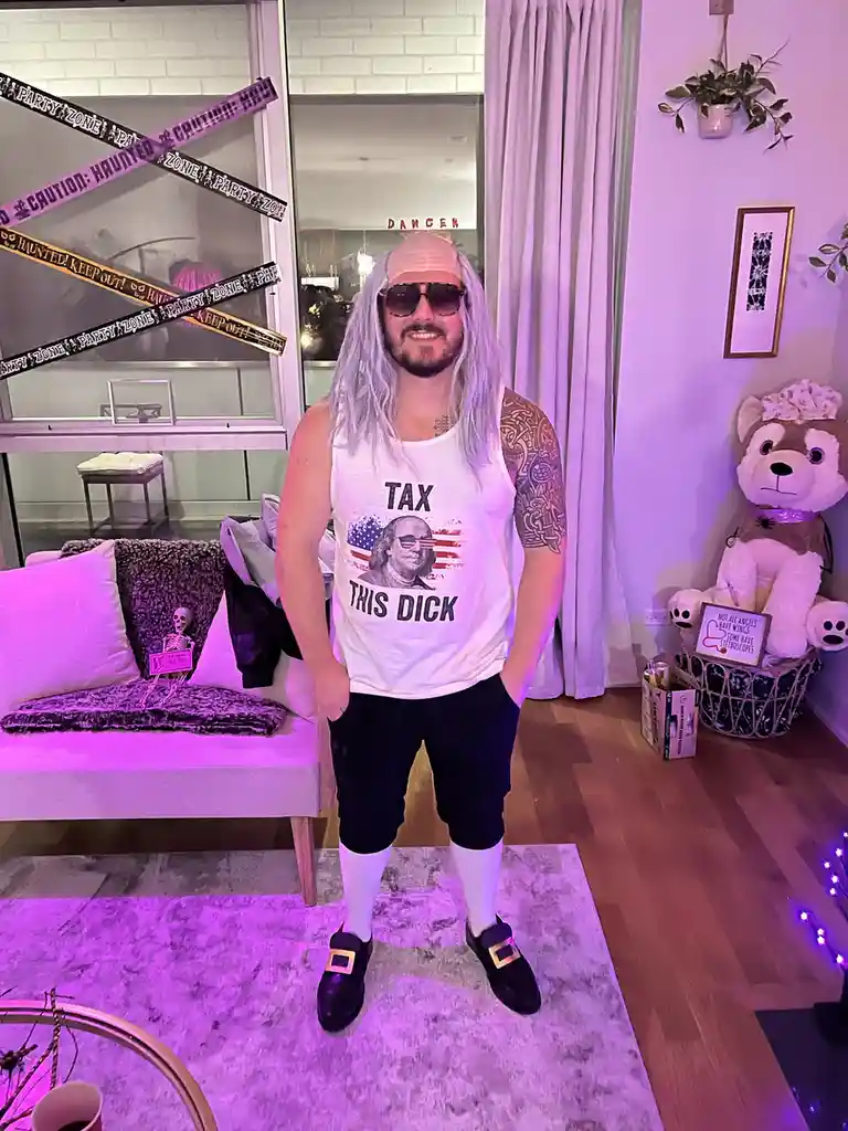 A man with a silver wig, sunglasses, and a white tank top featuring a graphic of Benjamin Franklin with the text 'TAX THIS…