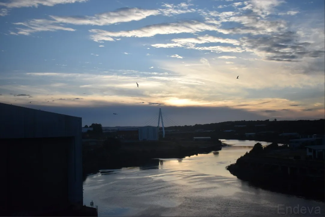 A serene sunset over a river with a modern cable-stayed bridge in the distance, silhouetted against a cloudy sky with birds…