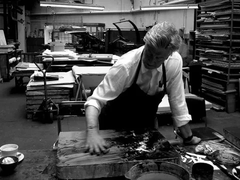 A black-and-white photo shows a person in a white shirt and dark apron, leaning over a large wooden block in what appears to…