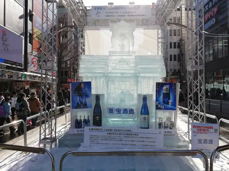 A large, intricate ice sculpture of a clock tower stands in a snowy urban plaza, surrounded by metal scaffolding and crowds…