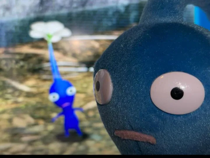 A close-up of a blue plush toy resembling a character from the video game 'Pikmin', with large, wide eyes and a downturned…