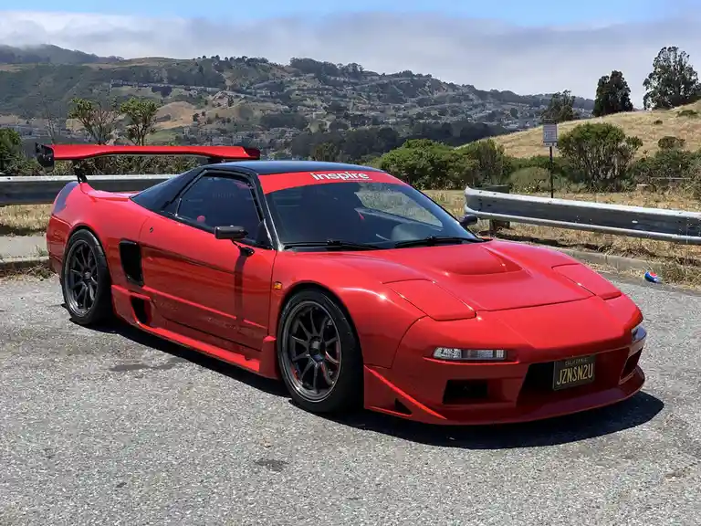 A red modified Honda NSX sports car with a large rear wing and 'inspire' decal on the windshield is parked on a roadside…