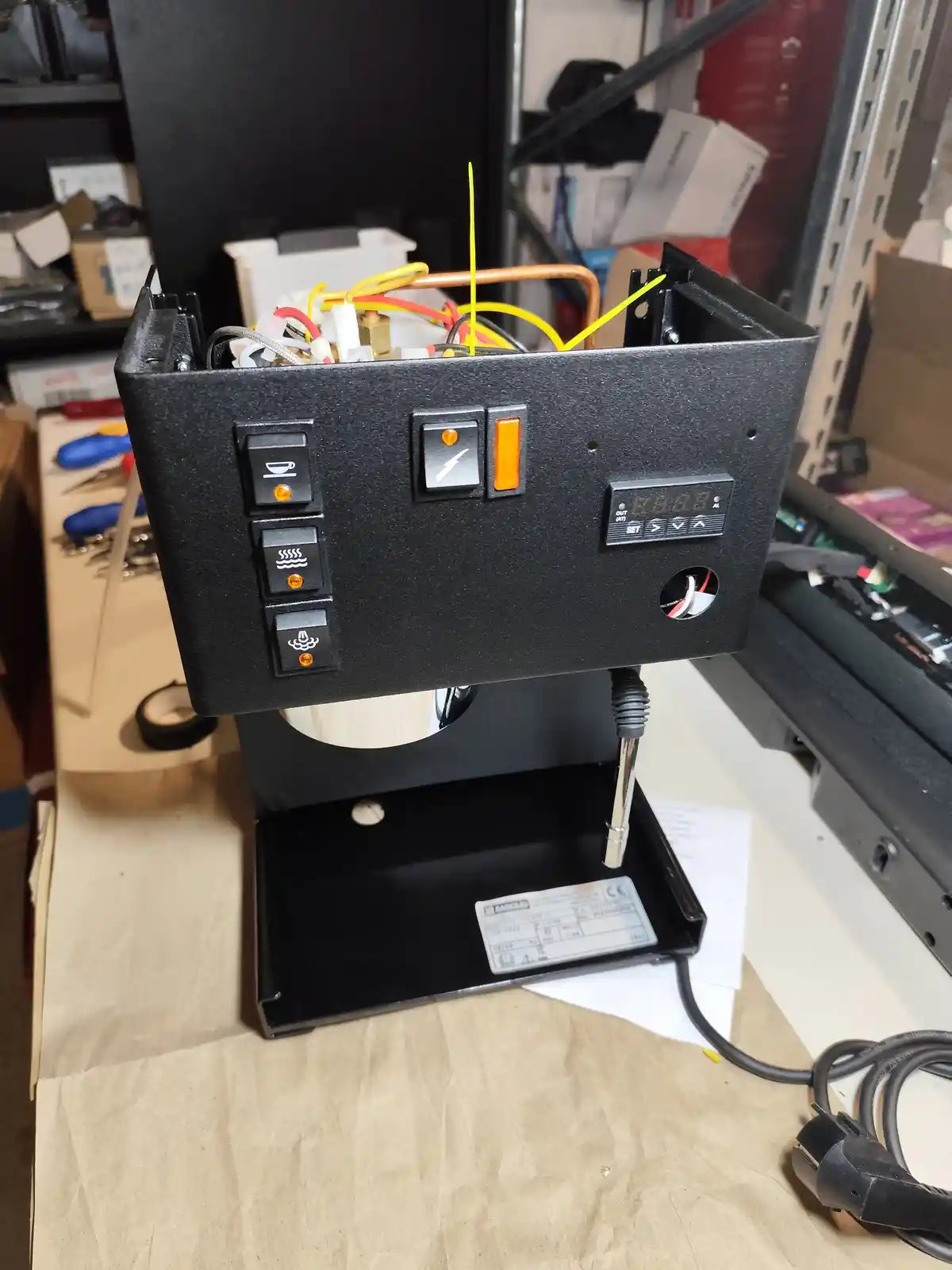 A black coffee machine with its top panel removed, exposing a tangle of colorful wires and copper tubing inside. The front…