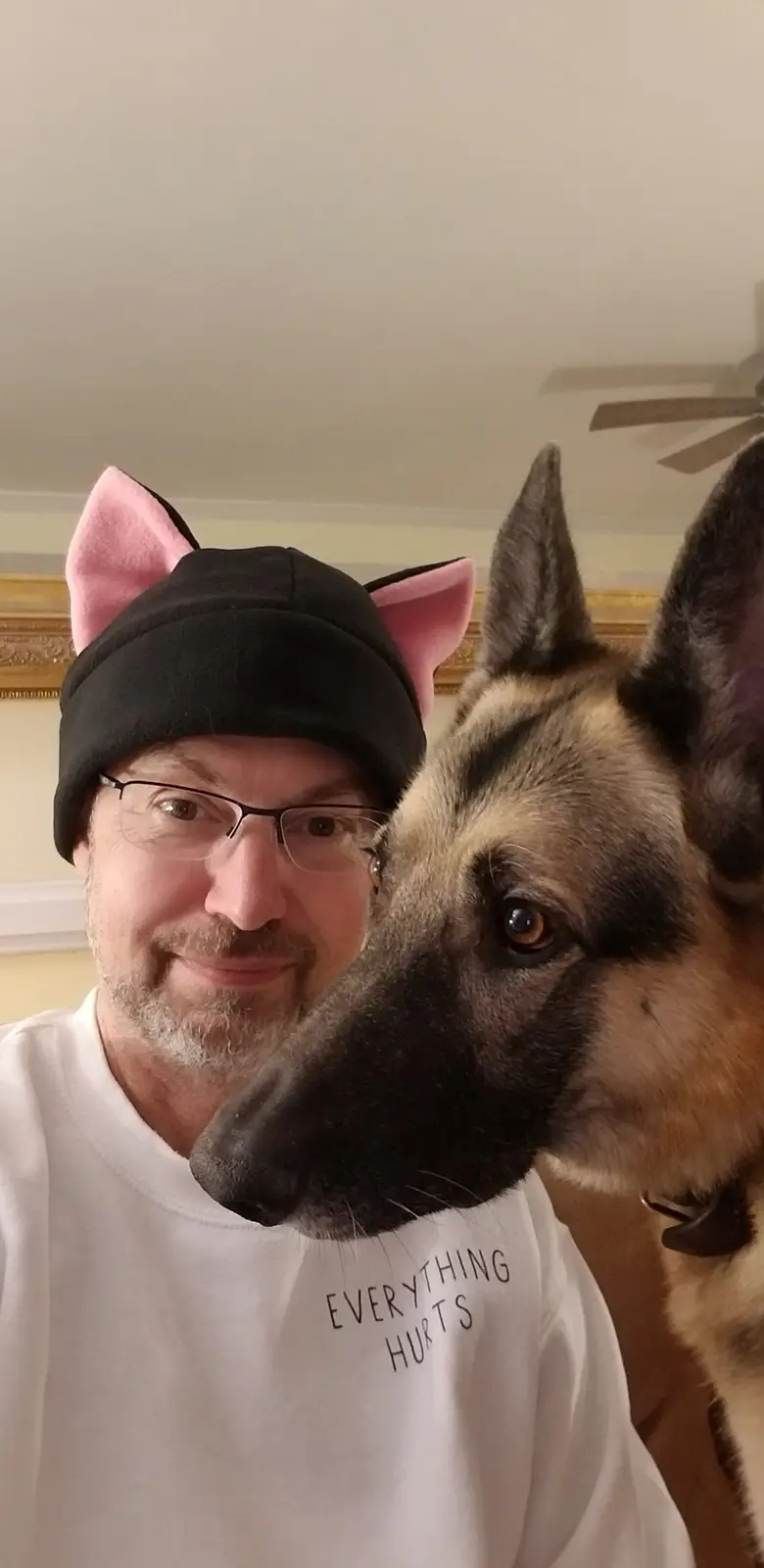 A man wearing a black beanie with pink cat ears and a white t-shirt that reads 'EVERYTHING HURTS' poses for a selfie with a…