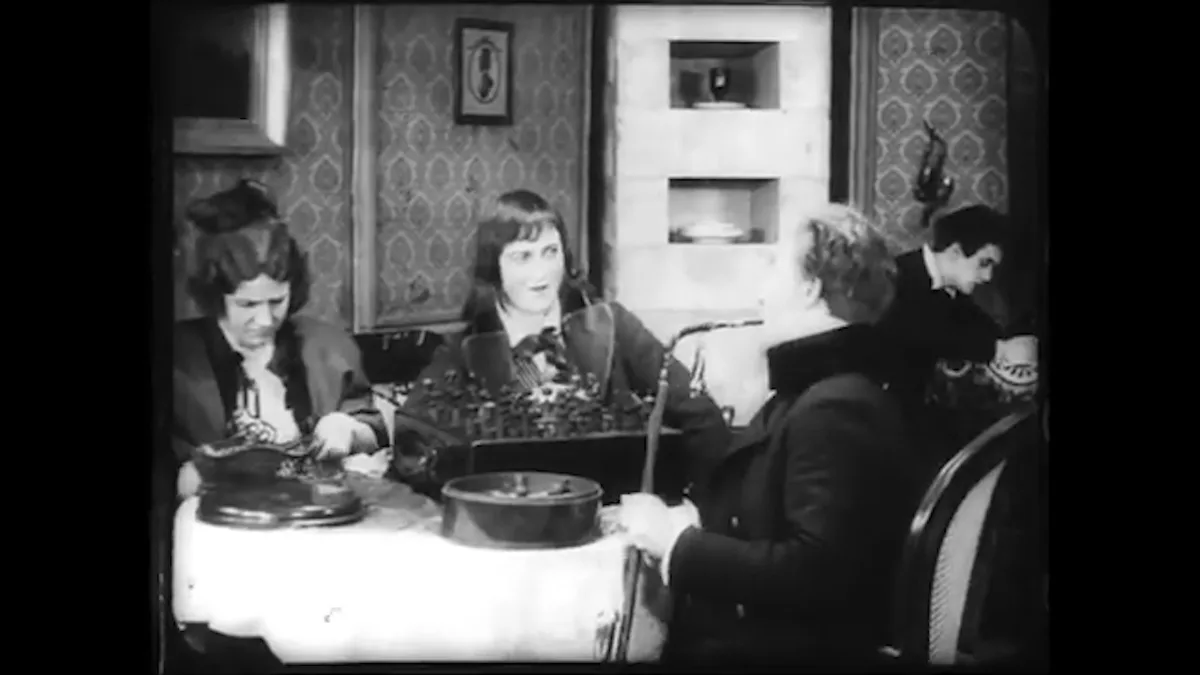 A black-and-white vintage film still shows four people seated around a table with old-fashioned typewriters. One woman in…