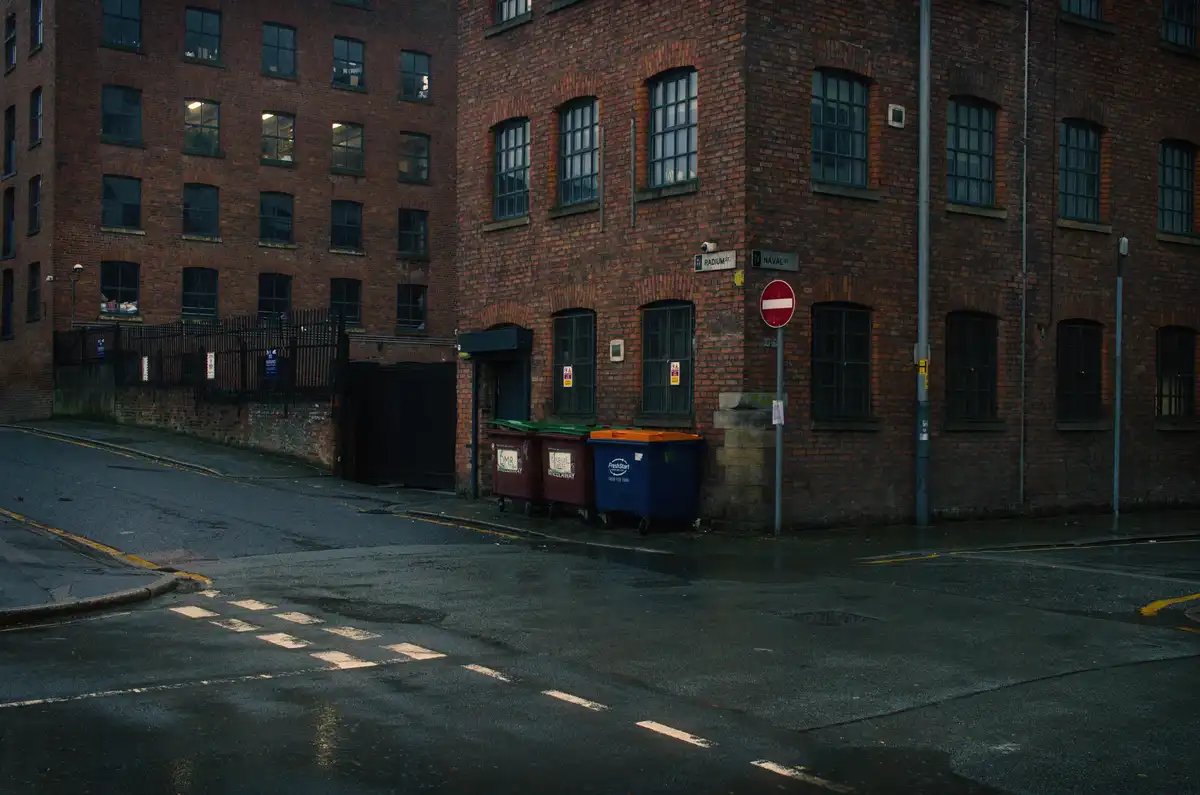 A wet, empty street corner with brick buildings, a 'no entry' sign, and three large trash bins. The overlaid text reads 'no…