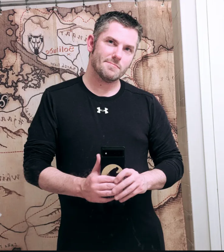 A man in a black Under Armour shirt stands in front of a shower curtain printed with a fantasy-style map, holding a phone…