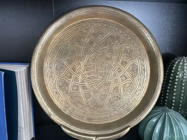 A close-up photo shows a large, ornate brass tray with intricate woven patterns, resting on a dark shelf next to a blue book…