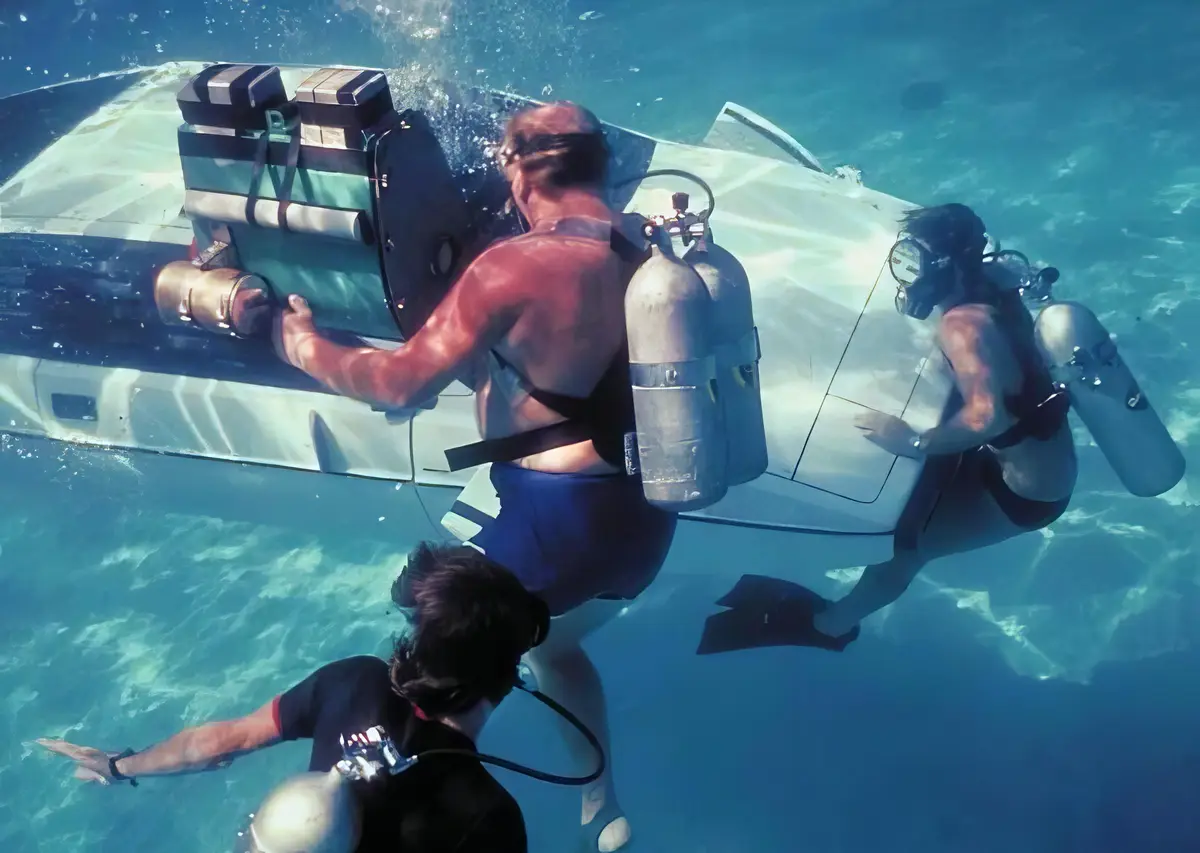 Three scuba divers are underwater next to a submerged white car, with one diver gripping the car door and another touching…