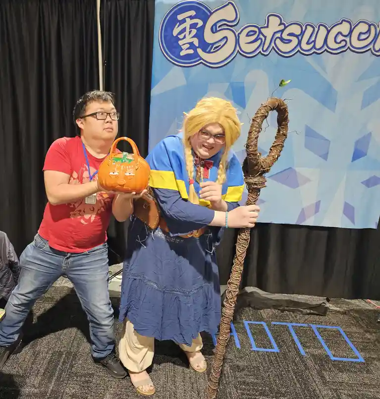 Two people pose at a convention with a 'Setsucon' backdrop. One person holds a carved pumpkin purse with a grumpy face…