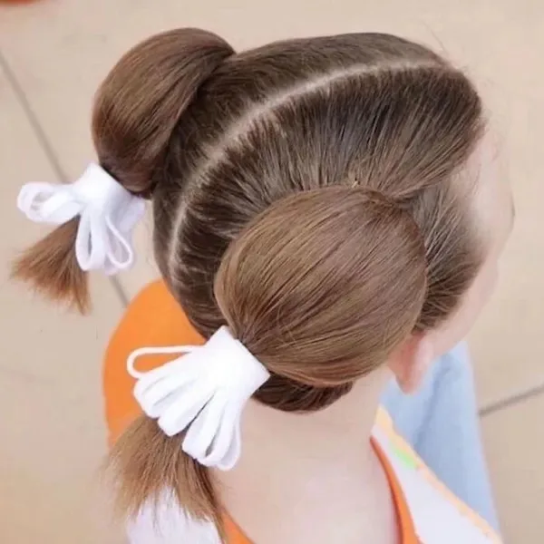 A close-up of the back of a child's head showing two neatly styled buns held by white fabric scrunchies, with the child…
