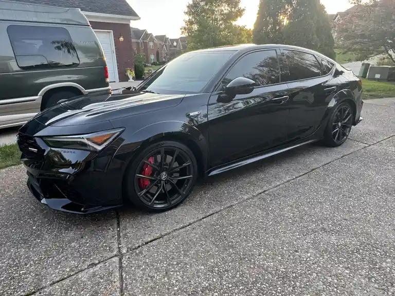 A glossy black Acura Integra Type S is parked in a driveway next to a green Ford Explorer, with a residential neighborhood…
